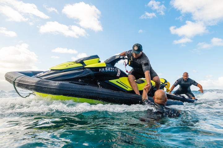 Men wearing Waihana Tropicam Black Short Sleeve Spring Suits conduct a waterman rescue training exercise on a Waihana-branded jet ski in open ocean waters.